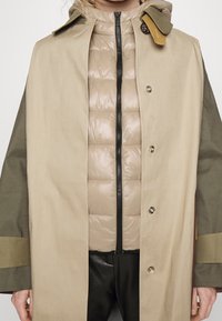 Beżowy i oliwkowozielony płaszcz z kapturem, z guzikami z przodu oraz pikowaną, beżową wewnętrzną kurtką z centralnym zamkiem błyskawicznym.