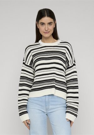 Frau mit langem dunklem Haar, die einen weiß-schwarz gestreiften Strickpullover und hellblaue Jeans trägt, vor einem einfarbigen Hintergrund stehend.