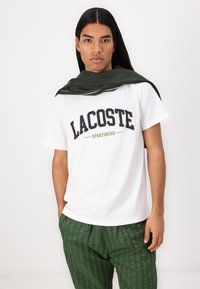 Biely bavlnený T-shirt s potlačou "LACOSTE SPORTSWEAR" v námorníckej a olivovej farbe. Model má na sebe tmavozelené vzorované nohavice a zelený šál prehodený cez plecia.