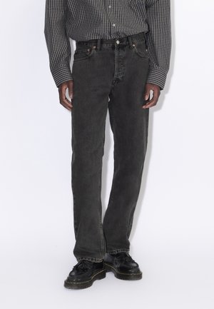 Persona con vaqueros negros desgastados, zapatos negros con cordones y una camisa de manga larga a cuadros en blanco y negro, de pie contra un fondo liso.