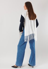 Scialle grigio chiaro con dettagli di frange indossato sopra un maglione blu navy; abbinato a jeans blu a vita alta e scarpe nere a punta.