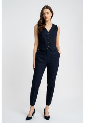 Kobieta o długich brązowych włosach ubrana w granatową bezrękawnikową kamizelkę na guziki, dopasowane spodnie oraz granatowe wysokie obcasy, stoi z jedną ręką w kieszeni.