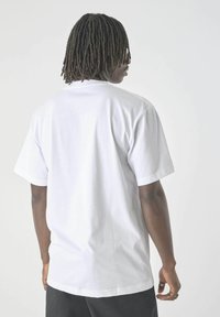 Person mit dunkler Haut und Dreadlocks, der ein einfaches weißes kurzärmliges T-Shirt und schwarze Hosen trägt, mit dem Rücken zu einem hellen Hintergrund.