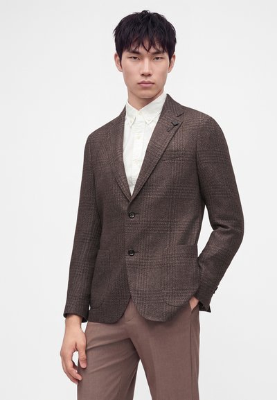 Jeune homme portant un blazer marron à carreaux sur une chemise blanche boutonnée et un pantalon marron, debout devant un fond clair uni.