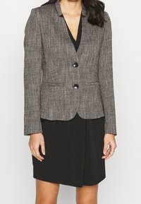 Blazer ajusté en tissu texturé gris et noir avec un motif pied-de-poule, deux boutons et des revers crantés, associé à une robe noire en dessous.