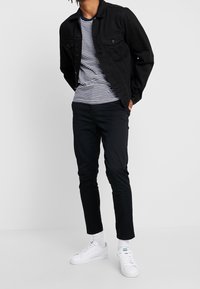 Giubbotto di jeans nero su una maglietta a righe, pantaloni neri e sneakers bianche. L'outfit presenta un design casual con colori e texture a contrasto.