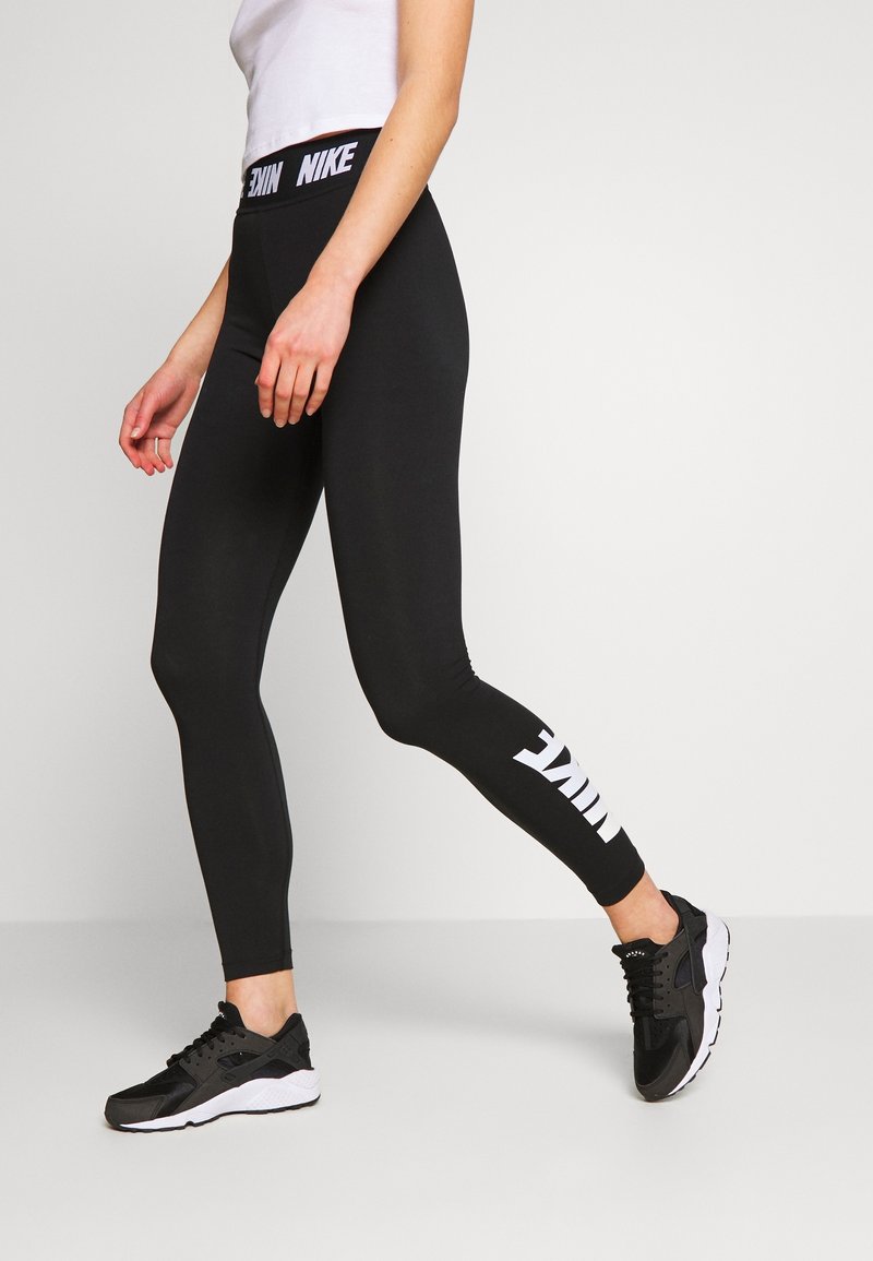 Leggings noirs avec un logo Nike blanc le long de la jambe inférieure et une ceinture de marque. Portés avec des chaussures de sport noires, montrant un design ajusté.
