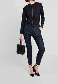 Camisa negra de manga larga con una cremallera dorada, combinada con pantalones ajustados azul marino. El modelo sostiene un pequeño bolso negro y lleva unos tacones puntiagudos negros.
