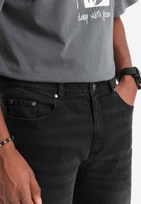Jeans in denim nero con vestibilità slim, dotati di una tasca frontale con rivetti metallici e chiusura a bottone. Abbinati a una maglietta grafica grigia.