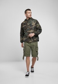 Camo-pullover in donkergroen en bruin, met een voorzak met rits; gecombineerd met olijfgroene cargoshorts en zwarte sneakers met witte veters.