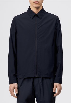 Mann iført en svart jakke med lange ermer, spiss krage og glidelås foran, kombinert med matchende svarte bukser.