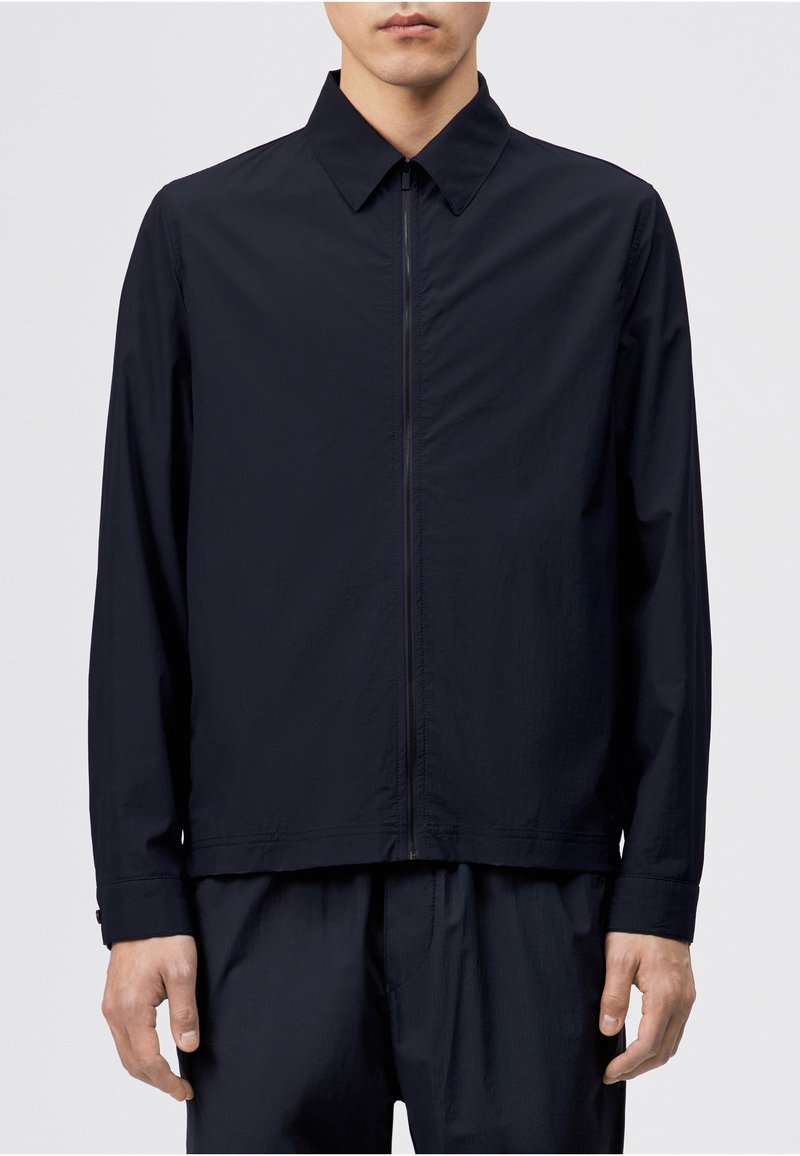 Homme portant une veste noire à manches longues avec un col pointu et une fermeture éclair à l'avant, associée à un pantalon noir assorti.