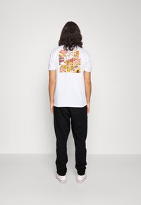 Camiseta blanca con un estampado gráfico abstracto y colorido en la parte posterior. Combinada con pantalones de chándal negros y zapatillas blancas.