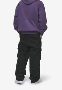 Fialová mikina s volným střihem spojená s volnými černými cargo kalhotami s bočními kapsami, doplněná bílými teniskami.