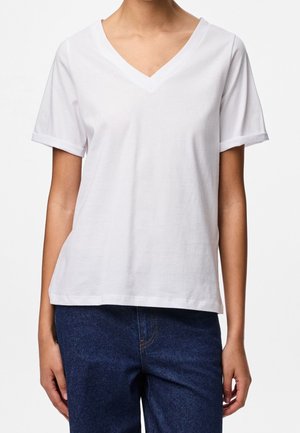 Personne portant un t-shirt blanc uni à col en V et manches courtes, et un jean bleu en denim, debout devant un fond blanc uni.
