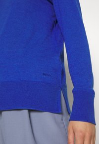 Gros plan d’une personne portant un pull bleu avec des poignets et un ourlet côtelés, associé à un pantalon gris clair, montrant un petit logo « BOSS ».