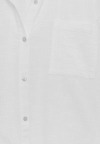 Chemise en lin blanche vue de face avec quatre boutons et une poche poitrine sur le côté gauche, montrant la texture et des détails de coutures subtiles.