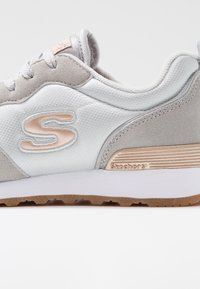 Gray and white sneaker with suede and mesh materials, featuring a textured side with a pink logo, white midsole, and brown rubber outsole.