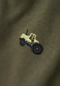 Diseño de tractor bordado en crema y navy sobre tejido verde oliva, con fina textura y detalles de costura.