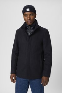 Cappotto in misto lana di colore blu navy con colletto alto e zip anteriore, abbinato a un berretto a coste scuro. Presenta tasche laterali e una vestibilità comoda.