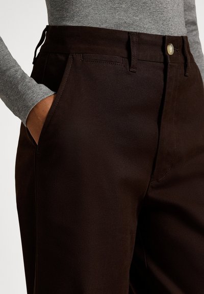 Άτομο που φοράει σκούρο καφέ παντελόνι με κουμπί και το χέρι στην τσέπη, συνδυασμένο με ανοιχτό γκρι μπλούζα μακρυμάνικη.