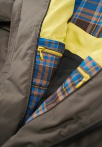 Gray jacket interior featuring yellow lining, blue and brown plaid pattern, and multiple zippered pockets with yellow hardware.