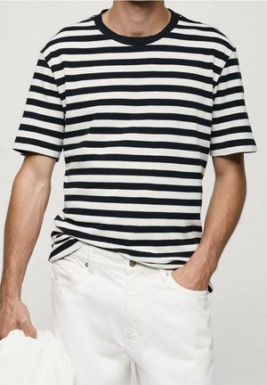 Mann trägt schwarz-weiß horizontal gestreiftes Kurzarmshirt und weiße Hose, eine Hand in der Tasche, hält ein weißes Kleidungsstück in der anderen Hand.