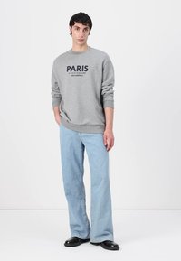 Felpa grigia con la scritta "PARIS" stampata in blu scuro, abbinata a jeans flare blu chiaro e scarpe nere, modello in posa su uno sfondo minimale.