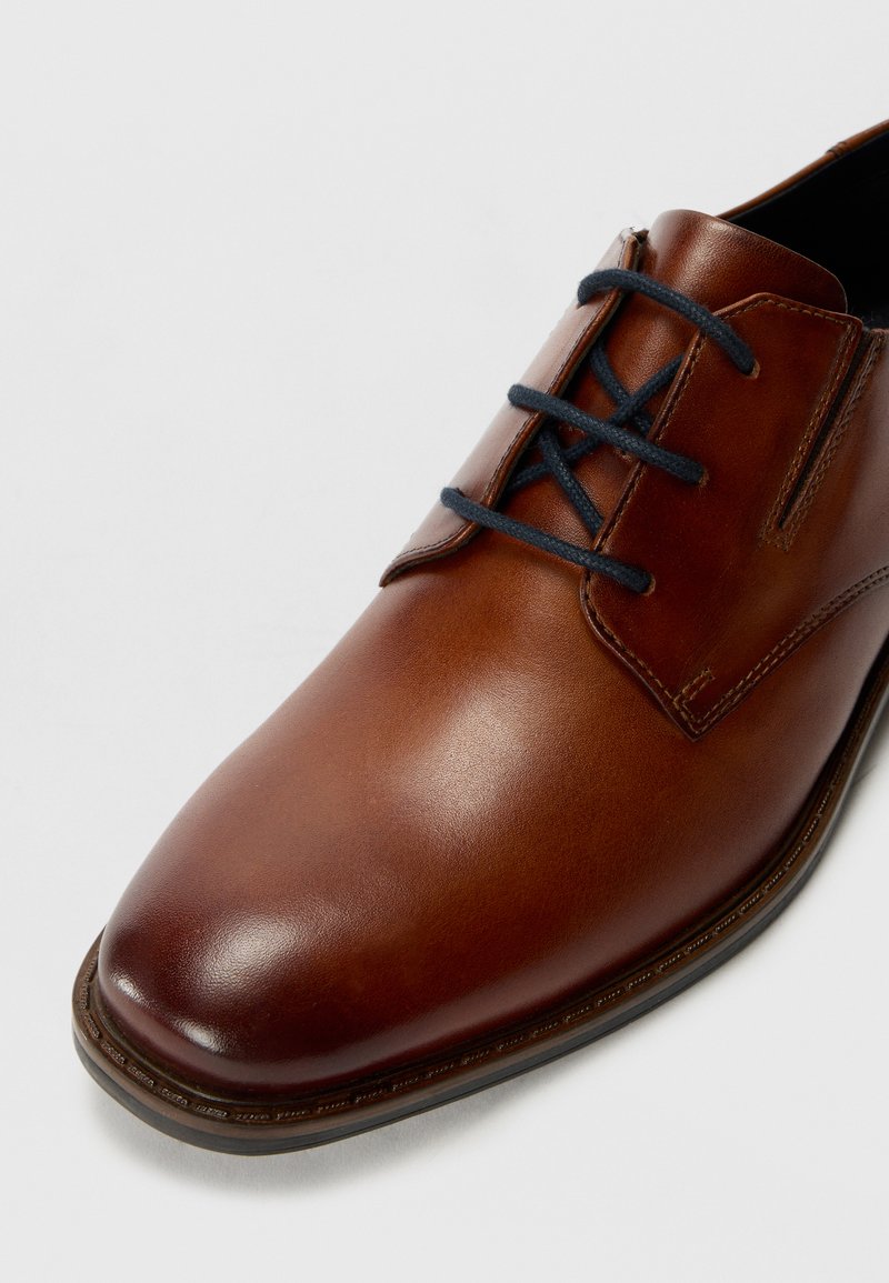 Zapato de cuero marrón con un acabado liso, punta redondeada y cordones en contraste de color navy. Presenta un diseño minimalista y detalles de costura sutiles.