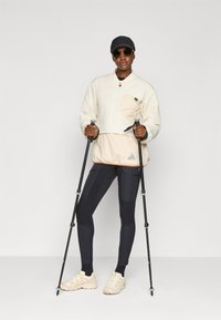 Világos bézs pulóver cipzáras zsebbe, fekete leggings, bézs cipők és fekete sapka. Túrabotokat használ. Simaszerkezetű anyag, minimalista design.