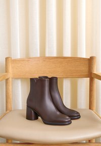 Paar dunkelbraune Leder-Stiefeletten mit Blockabsätzen auf einem beige gepolsterten Holzstuhl vor cremefarbenen Vorhängen.