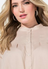 Blusa beige con colletto a fascia, apertura con bottoni e pieghe verticali. Tessuto liscio e leggero, impreziosito da una luce soffusa.