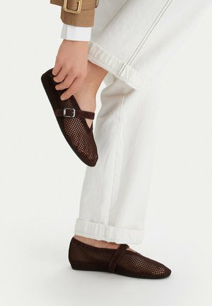 Osoba v bílých kalhotách drží jednu nohu zvednutou, ukazující tmavě hnědé síťované baleríny se sponami.