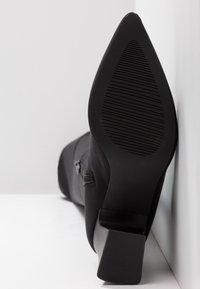 Botte noire à talon haut avec pointe effilée, tige en tissu lisse, fermeture éclair latérale et semelle en caoutchouc texturée avec des rainures verticales.