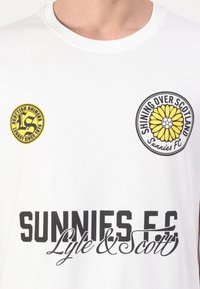 Tricou alb din bumbac, cu text negru "SUNNIES FC Lyle & Scott" și logo-uri circulare galbene: unul cu un floarea-soarelui, altul cu "Scottish Shiners."