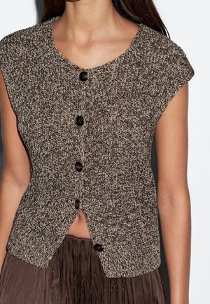 Cardigan sans manches en tricot brun et beige avec des boutons noirs, porté sur une jupe plissée foncée, montré sur une personne du cou jusqu'aux hanches.