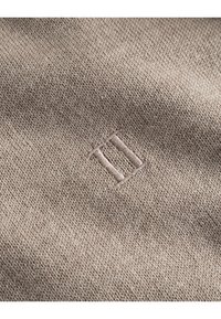 Beiger Strickstoff mit einem subtilen gestickten Logo, das aus zwei vertikalen Linien besteht. Weiche Textur mit einer glatten Oberfläche.