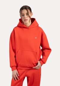 Felpa in cotone rossa con tasca frontale, polsini e orlo a coste, e un piccolo dettaglio con logo. Indossata con pantaloni della tuta rossi coordinati.
