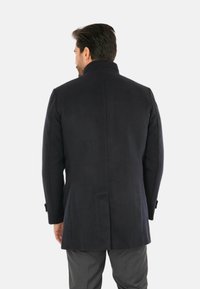 Cappotto in lana blu navy con colletto alto, spalle strutturate e schiena dritta. Presenta polsini con bottoni e un design pulito e minimalista.