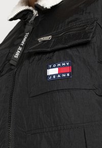 Czarna kurtka puchowa z zapinanym przodem, teksturowanym materiałem i kołnierzem wyłożonym Sherpą. Posiada haftowaną naszywkę z niebieskim i czerwonym logo.