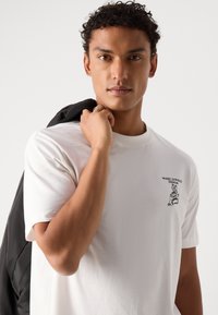 Witte katoenen t-shirt met een klein zwart grafisch ontwerp op de borst. Het model houdt een zwarte jas over één schouder.