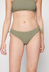 Bikini verde com padrão xadrez, apresentando laços laterais ajustáveis, um design justo e um tecido suave. A parte de cima tem um estilo cropped e bandeau.