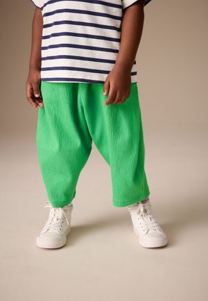 Barn iført grønne teksturerede vide bukser, navy- og hvidstribet skjorte og hvide snørebåndshøjtop-sneakers står på beige gulv.