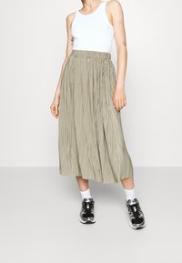 Person iført hvid ærmeløs top, beige plisseret midi nederdel, hvide sokker og sort-hvide sneakers foran en ensfarvet baggrund.