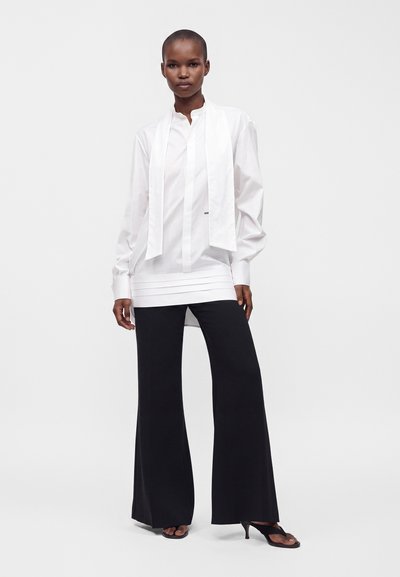 Femme debout portant une blouse blanche à manches longues avec des liens verticaux et un pantalon noir à jambes larges avec des sandales noires à talons.