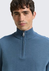 Blauer Zip-Kragen-Pullover mit strukturiertem Strickmuster, ausgestattet mit hohem Kragen und silbernem Reißverschluss. Der Stoff wirkt weich und leicht.
