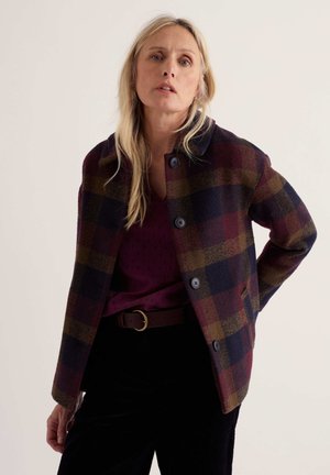 Femme aux longs cheveux blonds portant un manteau boutonné à carreaux sombre, une chemise bordeaux et un pantalon noir avec une ceinture marron, debout devant un fond uni.