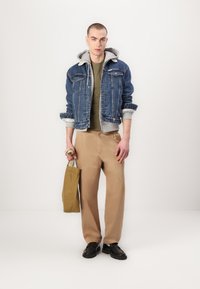 Denim jacket with gray hoodie underneath, olive green t-shirt, beige trousers, and black shoes. Holding a tan tote bag.