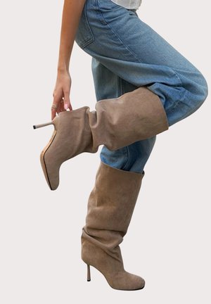 Personne portant un jean bleu et des bottes beige en daim souples montantes jusqu'au genou avec des talons aiguilles, levant une jambe et touchant la botte.
