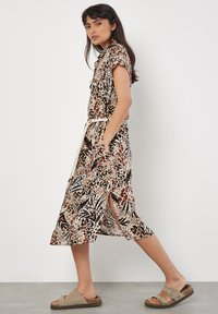 Vestito in tessuto con stampa floreale nei toni beige, marrone e nero. Maniche corte, cintura in vita, spacchi laterali e orlo al ginocchio. Indossato con sandali leggeri.